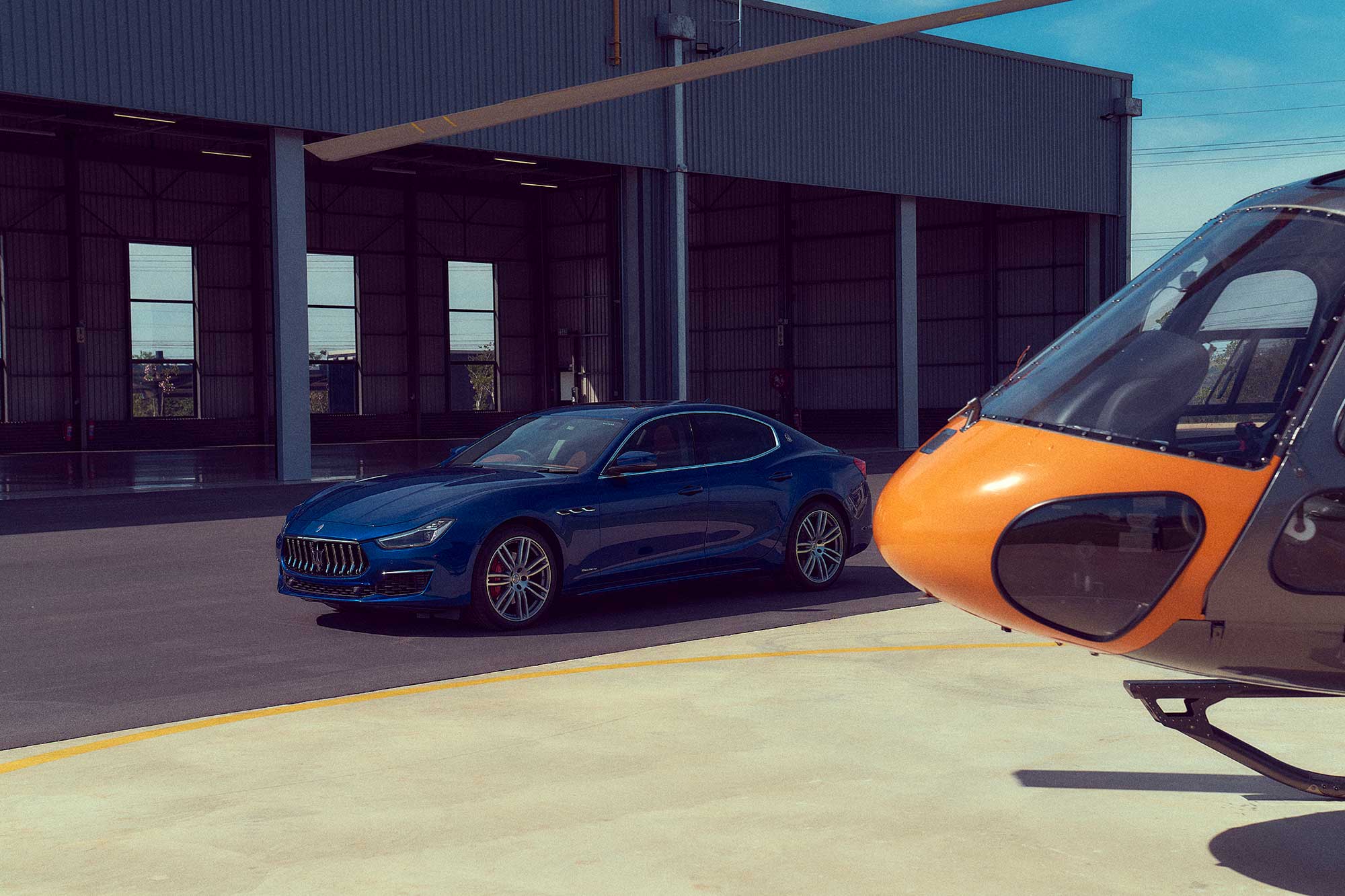 Stunning blue Maserati sports car parked in front of airplane hanger, showcasing its sleek design, aerodynamic lines, and iconic front grille. The sunlight reflects off the polished surface, highlighting its luxurious appeal. Capturing the energy and elegance of Maserati in Christchurch."